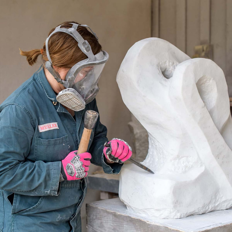 Elise Muller carving stone wearing respiratory protection, coveralls and pink work gloves.