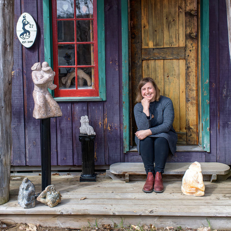 Elise Muller smiling and sitting on the steps of a purple wooden shed alongside 5 of her stone sculptures