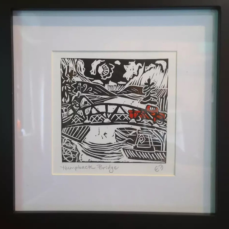 Framed square linocut print of a red truck crossing over a bridge with a river below and rolling hills and trees in the distance.