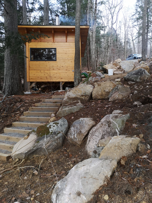 Small wooden cabin with black trim sits atop a hill with large rocks all around.