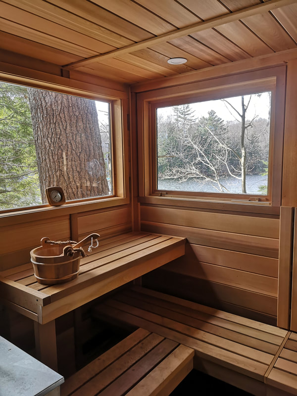 The inside of a beautiful wooden sauna with two large windows looking out on trees and water.